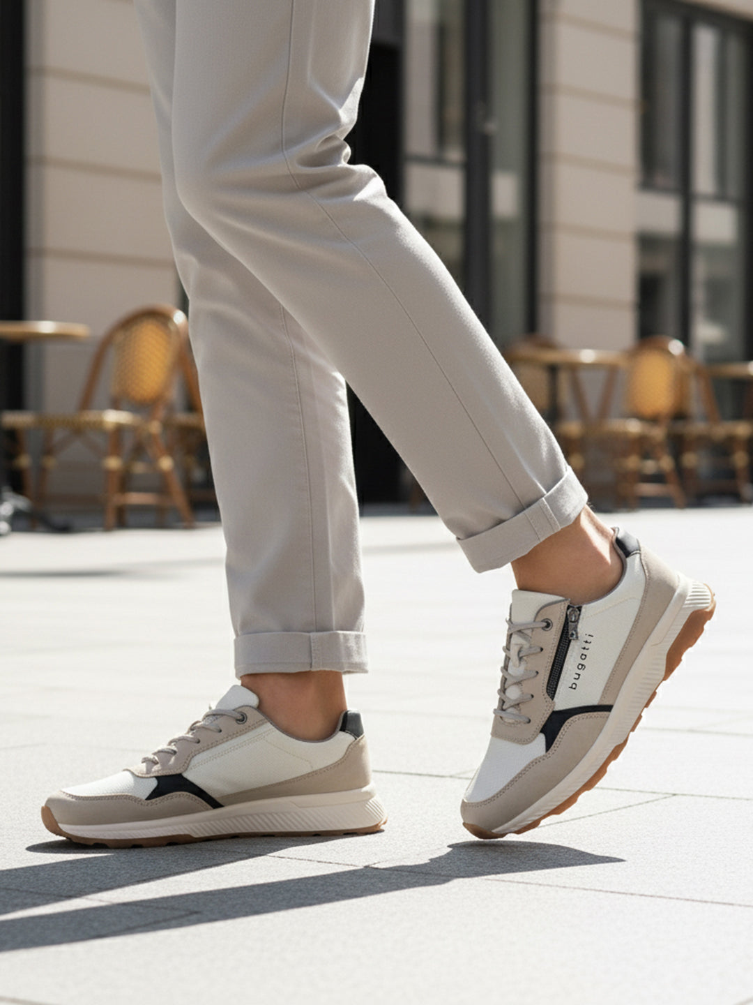 Lifestyle shot of bugatti Jerit Beige Comfort Sneakers, with zipper detail, worn with cuffed grey pants on a paved surface.
