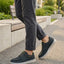 Lifestyle view of bugatti Fiero black comfort sneakers, worn with rolled dark gray pants, on a paved walkway.
