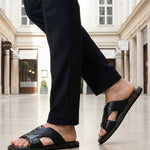 bugatti Cobie black leather sandals with a cross-strap design, paired with black trousers in a lifestyle shot.
