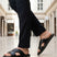 bugatti Cobie black leather sandals with a cross-strap design, paired with black trousers in a lifestyle shot.
