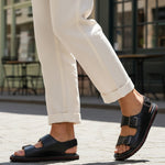Lifestyle shot of bugatti Cobie black leather sandals with comfort back strap, paired with white linen pants.
