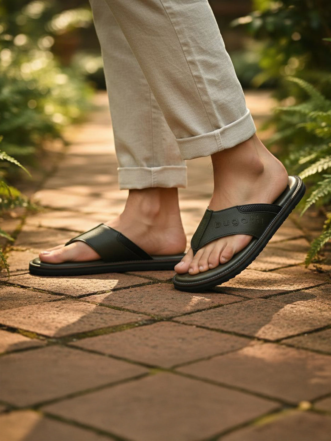 Lifestyle shot of bugatti Cobie dark green leather thong sandals, worn with rolled khaki pants, on a brick path.