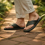 Lifestyle shot of bugatti Cobie dark green leather thong sandals, worn with rolled khaki pants, on a brick path.