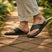 Lifestyle shot of bugatti Cobie dark green leather thong sandals, worn with rolled khaki pants, on a brick path.