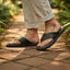 Lifestyle shot of bugatti Cobie dark green leather thong sandals, worn with rolled khaki pants, on a brick path.
