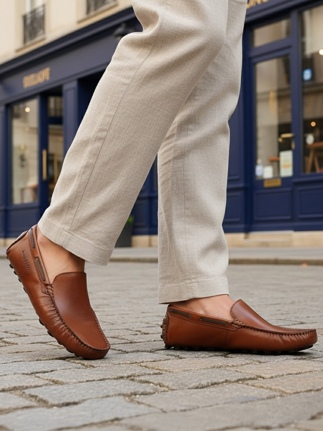 Lifestyle image of bugatti Manfield cognac leather slip-on shoes with visible textured beige pants on a cobblestone street.