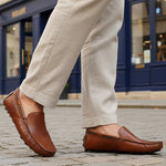 Lifestyle image of bugatti Manfield cognac leather slip-on shoes with visible textured beige pants on a cobblestone street.