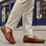 Lifestyle image of bugatti Manfield cognac leather slip-on shoes with visible textured beige pants on a cobblestone street.