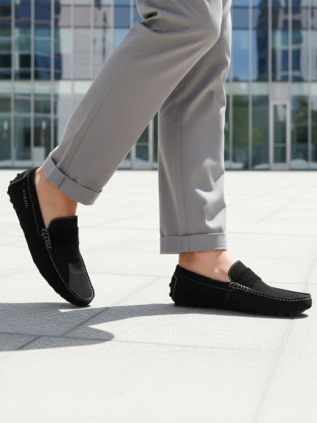Lifestyle shot of a bugatti Manfield black leather slip-on shoe, paired with gray casual pants.
