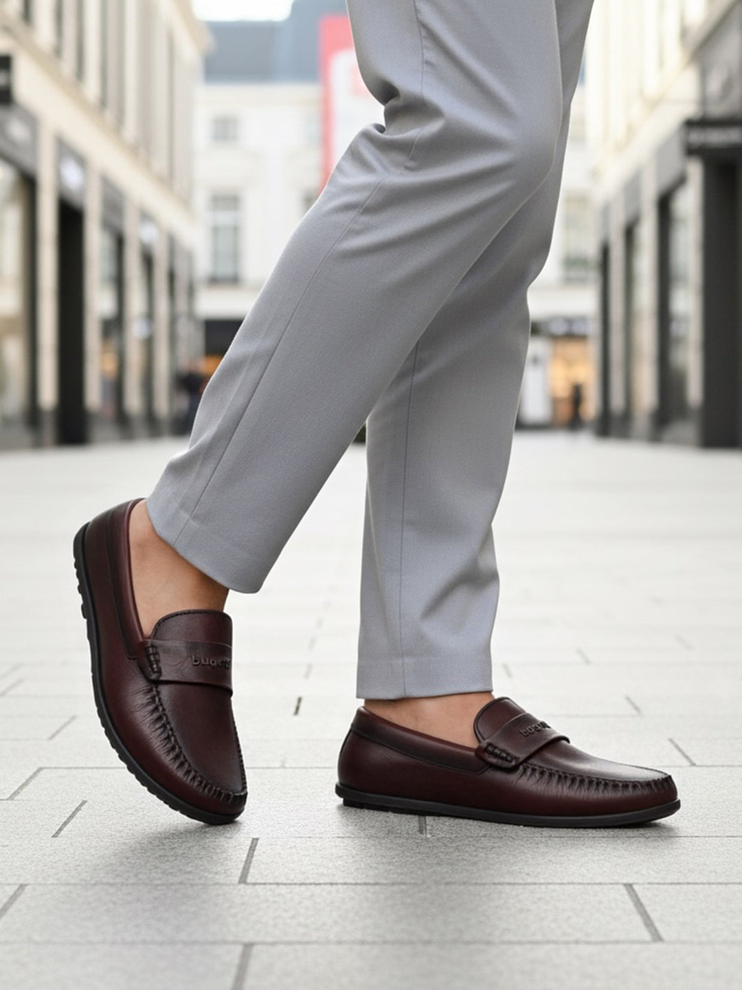 Lifestyle shot of a bugatti Linex slip-on shoe in mid-brown leather, paired with gray slacks and worn on a tiled street.
