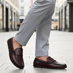 Lifestyle shot of a bugatti Linex slip-on shoe in mid-brown leather, paired with gray slacks and worn on a tiled street.
