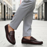 Lifestyle shot of a bugatti Linex slip-on shoe in mid-brown leather, paired with gray slacks and worn on a tiled street.
