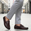 Lifestyle shot of a bugatti Linex slip-on shoe in mid-brown leather, paired with gray slacks and worn on a tiled street.
