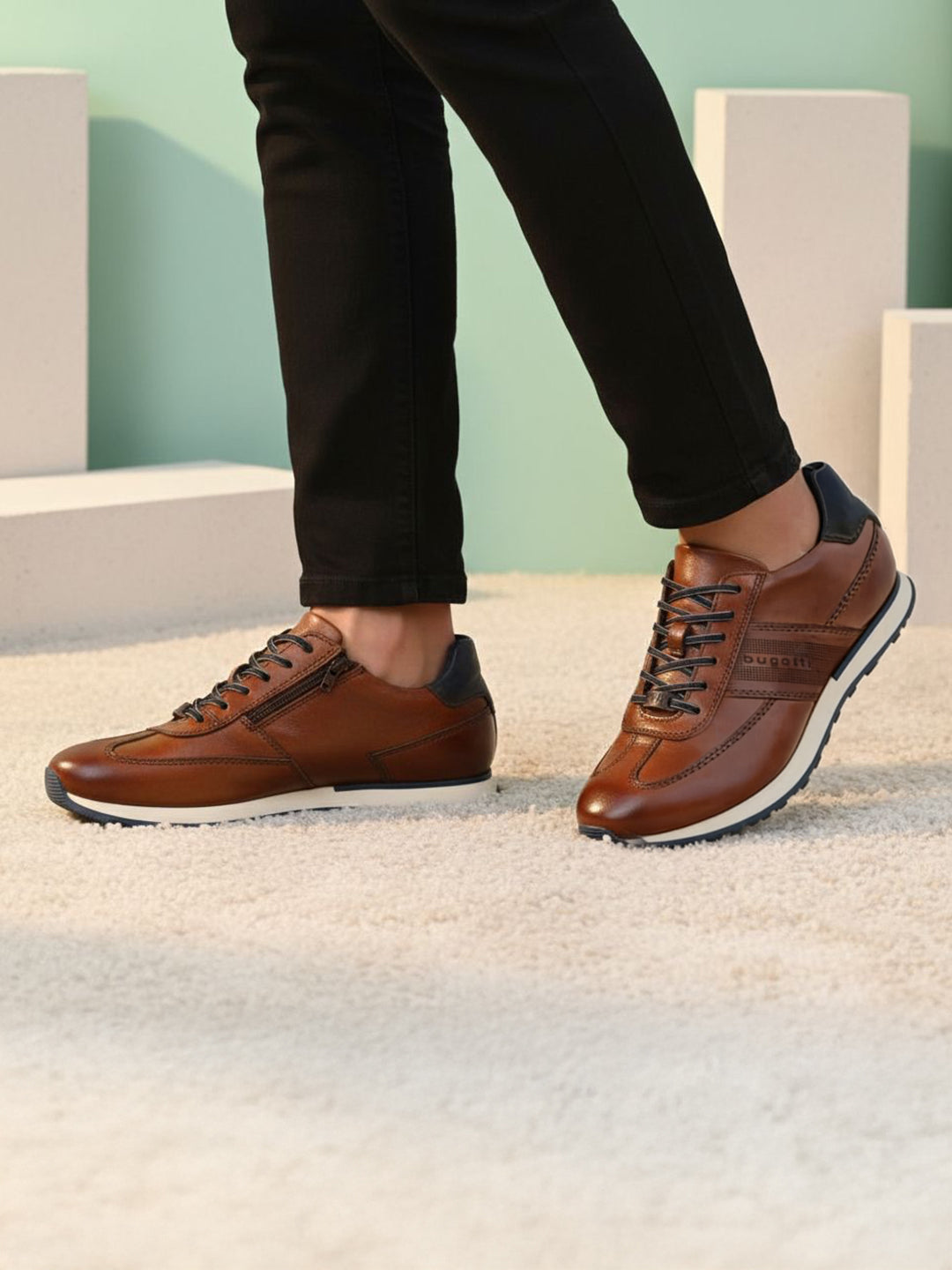 Lifestyle shot of brown leather bugatti sneakers with black laces and dark blue accent, worn with black pants.
