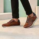 Lifestyle shot of brown leather bugatti sneakers with black laces and dark blue accent, worn with black pants.
