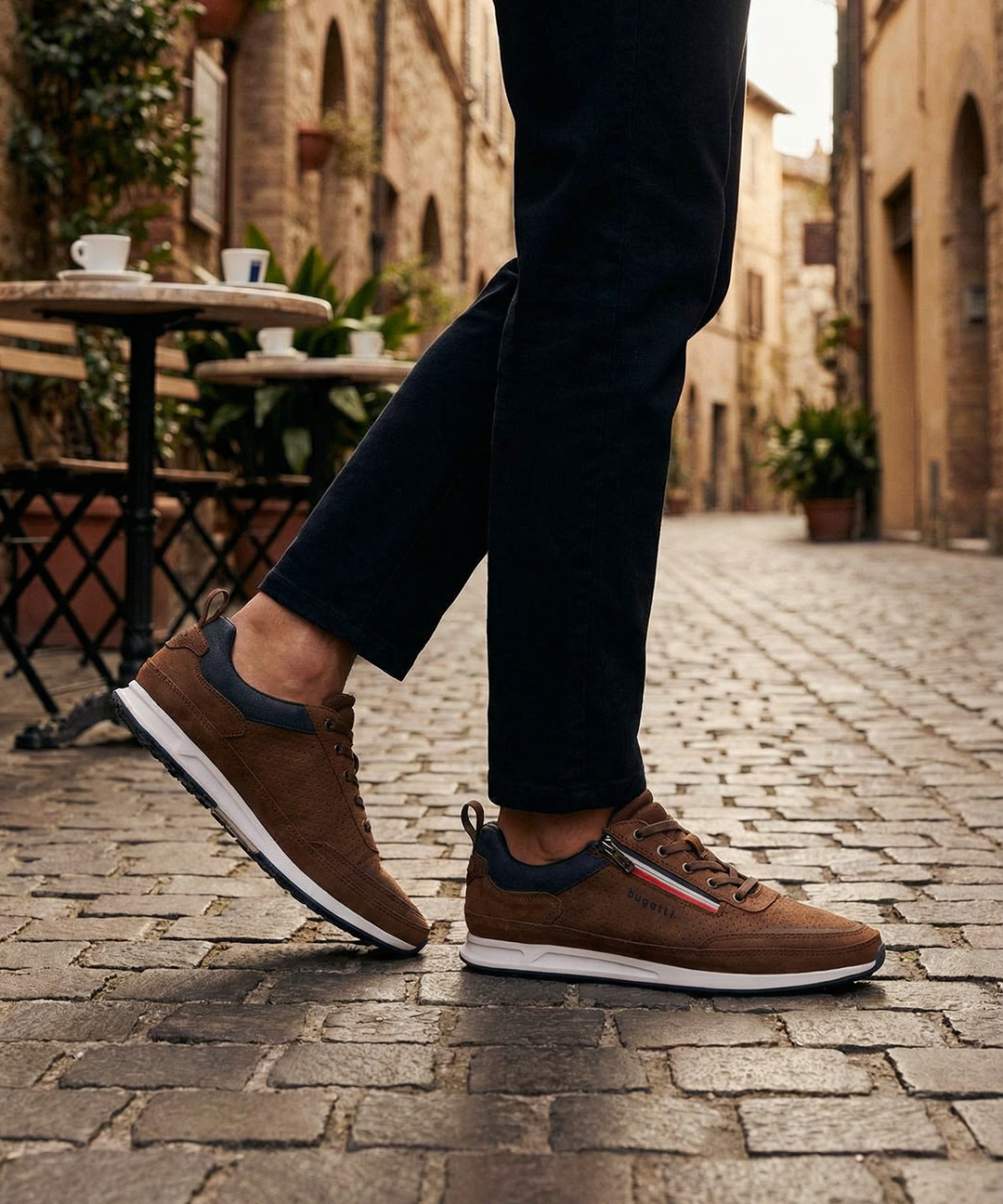 Lifestyle side view of the bugatti Ross Cognac Comfort Mid Top Sneakers, with blue accent and zipper detail, worn with black pants.
