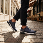 Dark blue sneakers by bugatti with gray accents and white soles, worn with black trousers on a cobblestone street.
