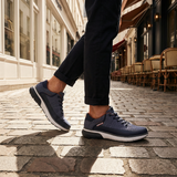 Dark blue sneakers by bugatti with gray accents and white soles, worn with black trousers on a cobblestone street.
