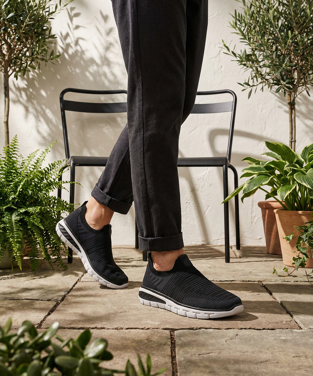 Lifestyle image of bugatti Yucatan black comfort sneakers, with white soles, worn with cuffed black pants.
